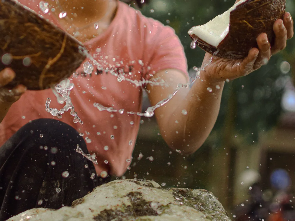Hands cracking open a fresh coconut, water splashing in the light